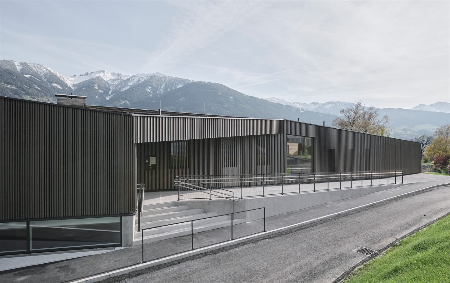 Kindergarten und Kinderkrippe Vomp — dunkel verschalter Baukörper vor verschneiter Tiroler Bergkette